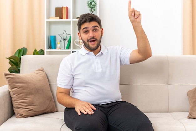 Jeune homme en vêtements décontractés à l'air surpris et heureux montrant l'index ayant une nouvelle idée assis sur un canapé dans un salon lumineux