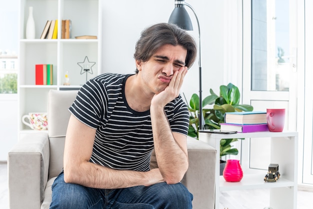 Jeune homme en vêtements décontractés assis sur la chaise avec une expression triste sur le visage penché la tête sur le bras dans un salon lumineux