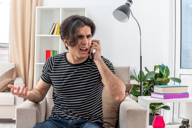 Jeune homme en vêtements décontractés ayant l'air confus et mécontent tout en parlant au téléphone portable assis sur la chaise dans un salon lumineux