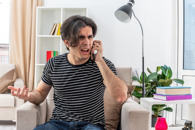 Jeune homme en vêtements décontractés ayant l'air confus et mécontent tout en parlant au téléphone portable assis sur la chaise dans un salon lumineux