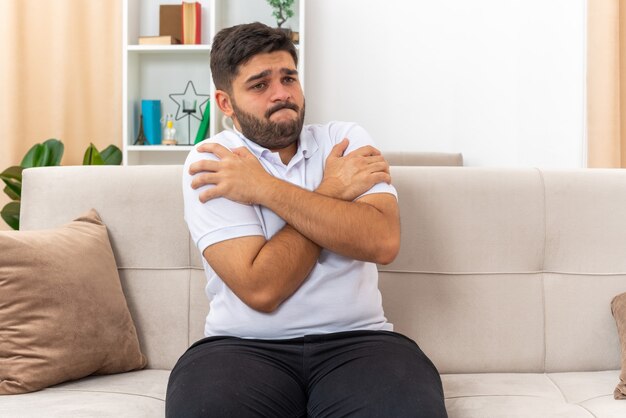 Jeune homme en vêtements décontractés à côté confus et inquiet avec les bras croisés sur la poitrine assis sur un canapé dans un salon lumineux