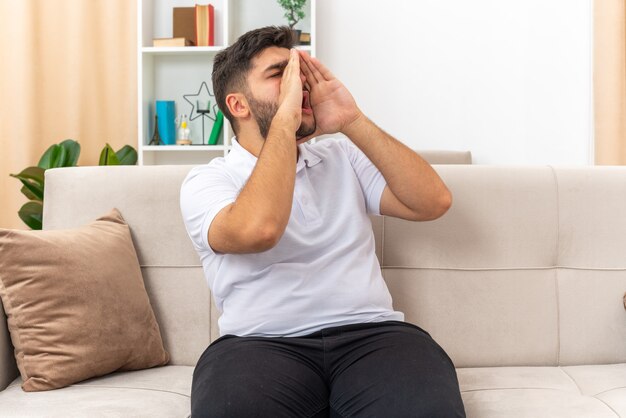 Jeune homme en vêtements décontractés criant ou appelant avec les mains près de la bouche assis sur un canapé dans un salon lumineux