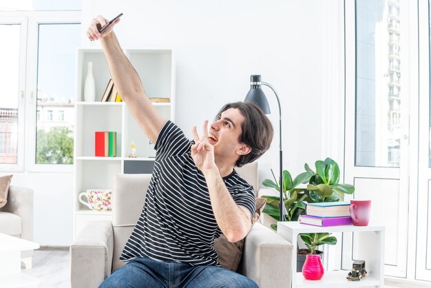 Jeune homme en vêtements décontractés faisant selfie à l'aide d'un smartphone heureux et positif montrant un signe v assis sur la chaise dans un salon lumineux