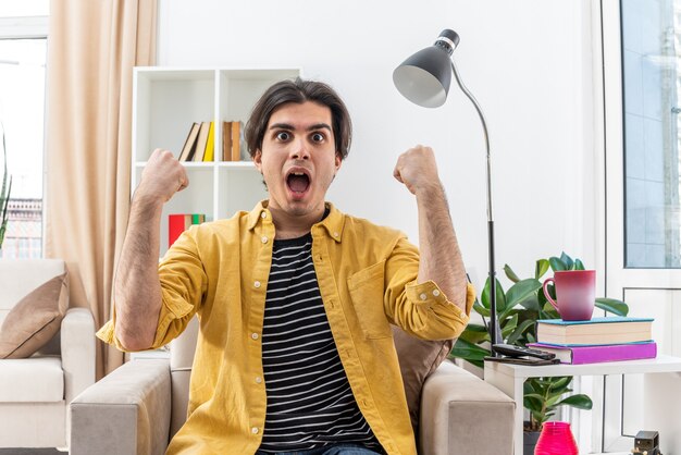 Jeune homme en vêtements décontractés, heureux et excité, serrant les poings assis sur la chaise dans un salon lumineux