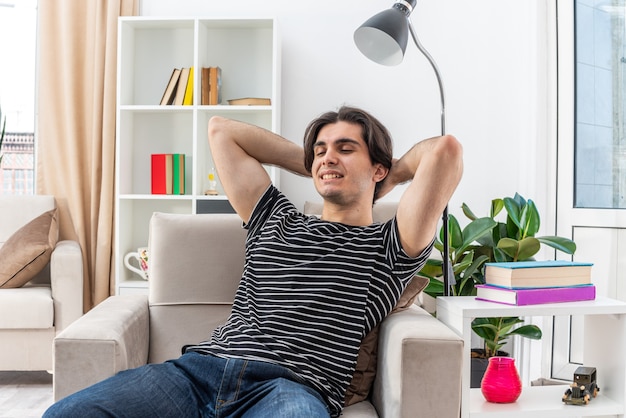 Jeune homme en vêtements décontractés, heureux et positif, passer un week-end de détente assis sur la chaise dans un salon lumineux