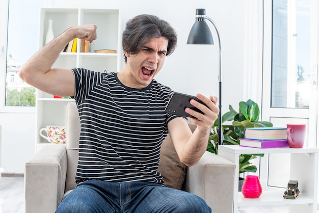 Jeune homme en vêtements décontractés jouant à des jeux à l'aide d'un smartphone heureux et excité levant le poing comme un gagnant assis sur la chaise dans un salon lumineux