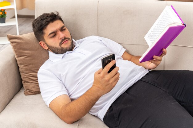 Jeune homme en vêtements décontractés avec un livre tenant un smartphone le regardant avec un visage sérieux passant le week-end à la maison allongé sur un canapé dans un salon lumineux