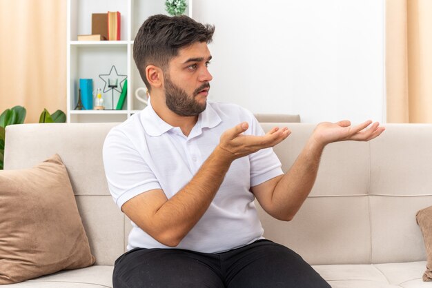 Jeune homme en vêtements décontractés regardant de côté confus présentant avec des bras quelque chose assis sur un canapé dans un salon lumineux