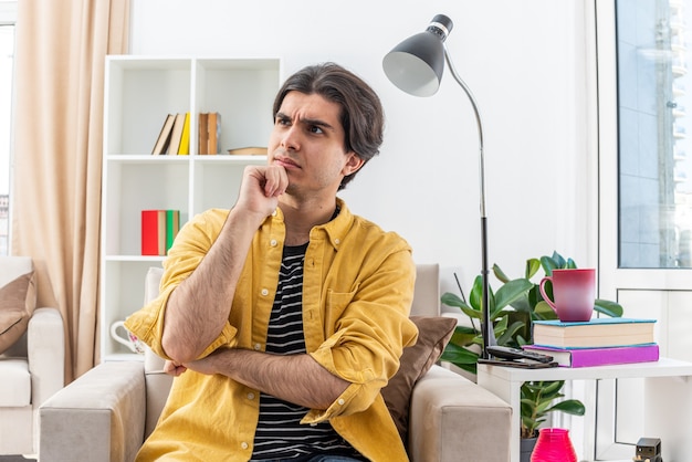 Jeune homme en vêtements décontractés regardant de côté avec une expression pensive avec la main sur le menton pensant assis sur la chaise dans un salon lumineux