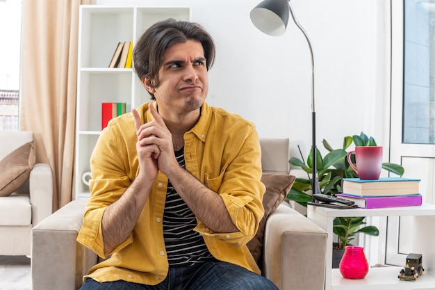 Jeune homme en vêtements décontractés regardant de côté avec une expression pensive pensant assis sur la chaise dans un salon lumineux