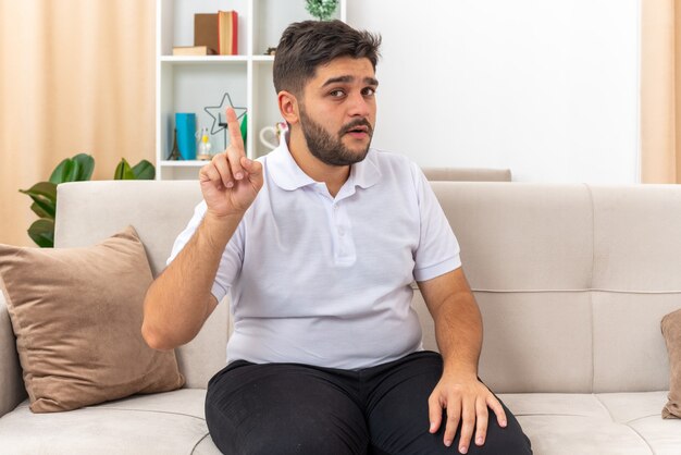 Jeune homme en vêtements décontractés regardant montrant l'index ayant une nouvelle idée assis sur un canapé dans un salon lumineux