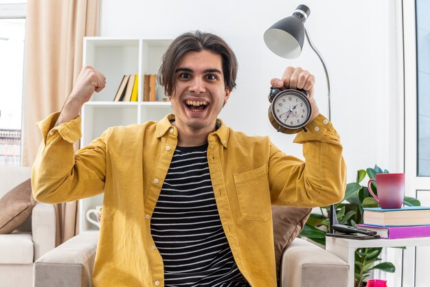 Jeune homme en vêtements décontractés tenant un réveil heureux et excité levant le poing comme un vigneron assis sur la chaise dans un salon lumineux