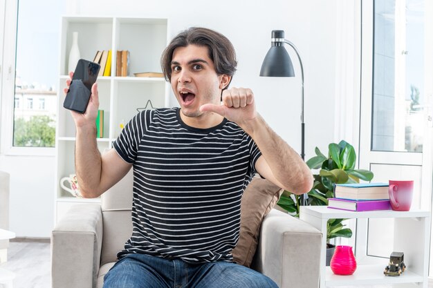 Jeune homme en vêtements décontractés tenant un smartphone à l'air heureux et excité montrant les pouces vers le haut assis sur la chaise dans un salon lumineux