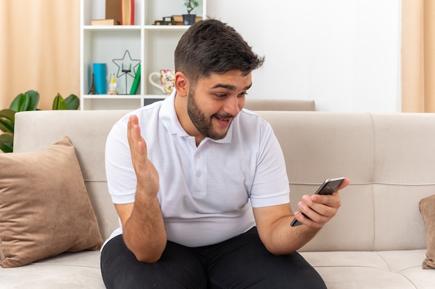 Jeune homme en vêtements décontractés tenant un smartphone le regardant heureux et excité assis sur un canapé dans un salon lumineux