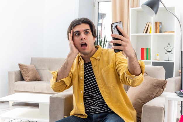 Jeune homme en vêtements décontractés tenant un smartphone le regardant inquiet et confus assis sur la chaise dans un salon lumineux