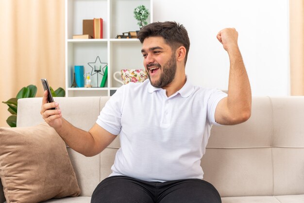 Jeune homme en vêtements décontractés tenant un smartphone serrant le poing heureux et excité assis sur un canapé dans un salon lumineux