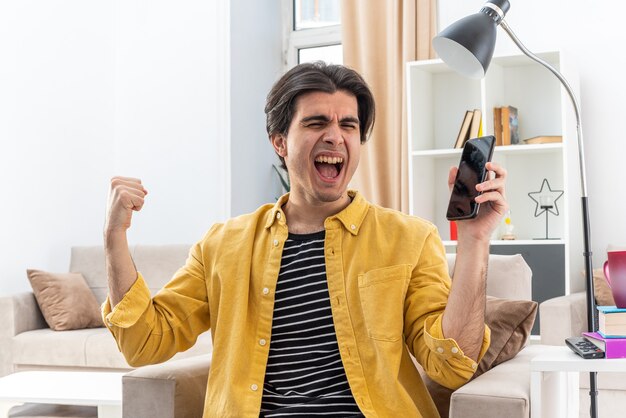 Jeune homme en vêtements décontractés tenant un smartphone serrant le poing heureux et excité se réjouissant de son succès assis sur la chaise dans un salon lumineux