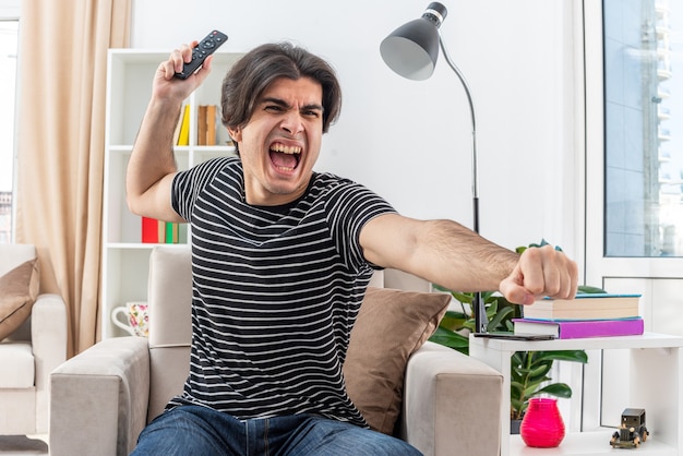 Jeune homme en vêtements décontractés tenant la télécommande de la télévision en criant se déchaîner assis sur la chaise dans un salon lumineux