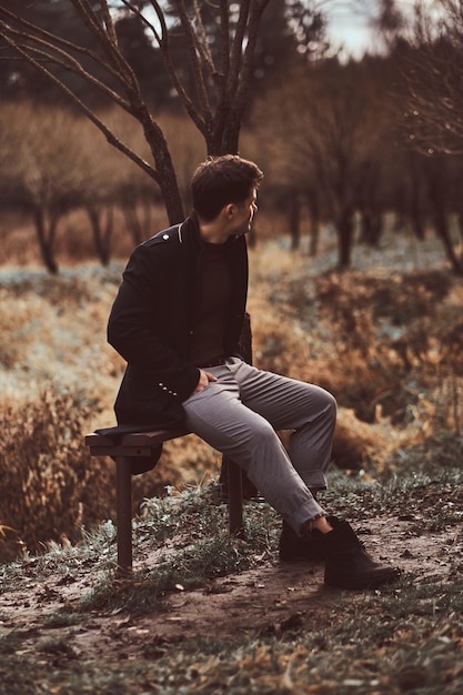 Photo gratuite un jeune homme vêtu d'un manteau noir assis sur un banc dans le bosquet de la forêt d'automne le soir.