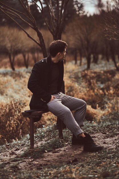 Un jeune homme vêtu d'un manteau noir assis sur un banc dans le bosquet de la forêt d'automne le soir.