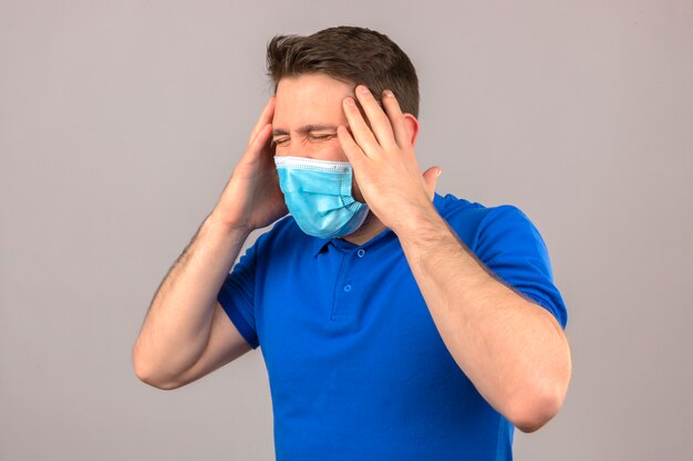 Jeune homme vêtu d'un polo bleu dans un masque de protection médicale à la recherche de mal et malade debout avec la main sur la tête souffrant de maux de tête sur un mur blanc isolé