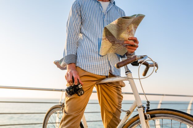 Jeune homme voyageant à vélo au bord de la mer en vacances d'été au bord de la mer au coucher du soleil, tenant une carte touristique avec appareil photo