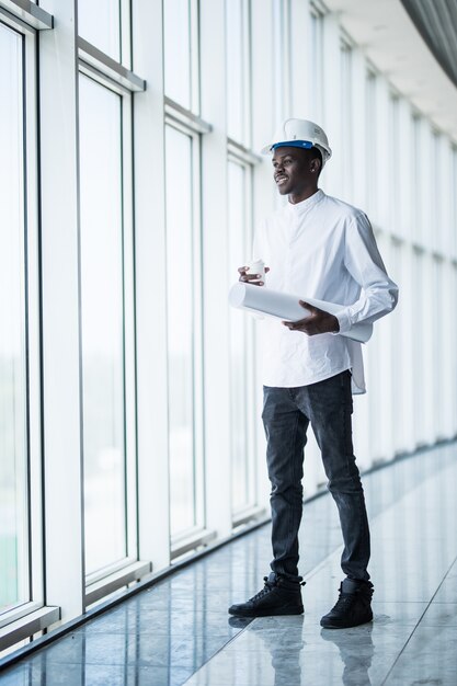 Jeune ingénieur afro-américain avec des imprimés bleus devant des fenêtres panoramiques au bureau