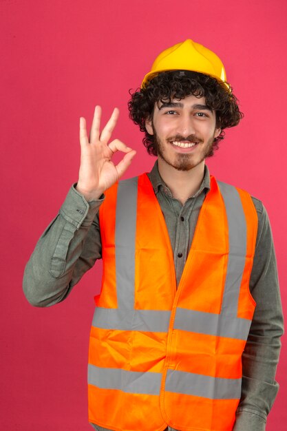 Jeune ingénieur beau barbu portant un casque de sécurité et gilet souriant faisant signe ok debout sur un mur rose isolé
