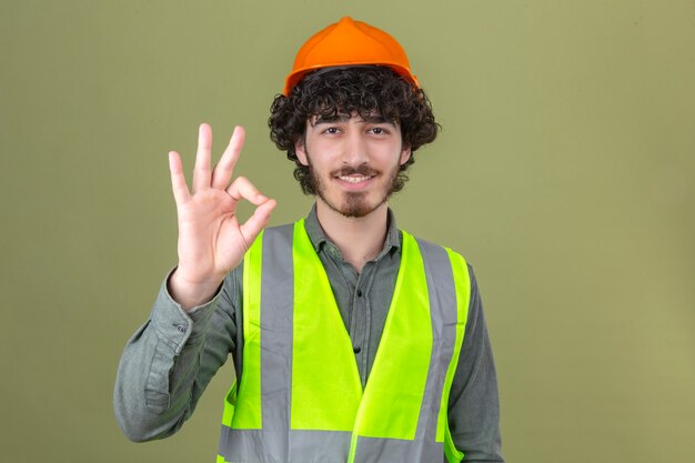 Jeune ingénieur beau barbu portant un casque de sécurité et gilet souriant faisant signe ok debout sur mur vert isolé