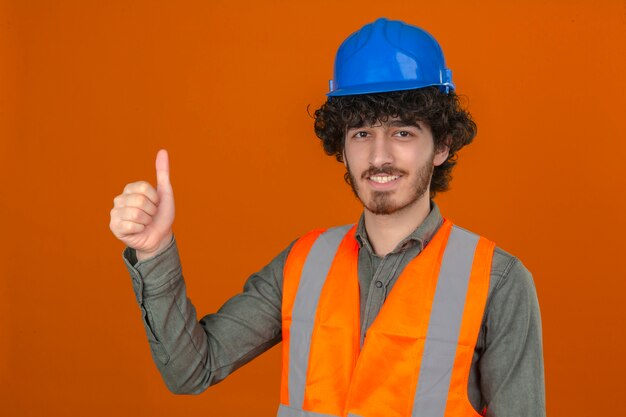 Jeune ingénieur beau barbu portant un casque de sécurité et un gilet avec sourire sur le visage montrant le pouce vers le haut sur un mur orange isolé