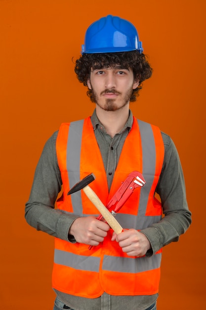 Photo gratuite jeune ingénieur beau barbu portant un casque de sécurité et un gilet tenant une clé à molette et un marteau avec un visage sérieux debout sur un mur orange isolé