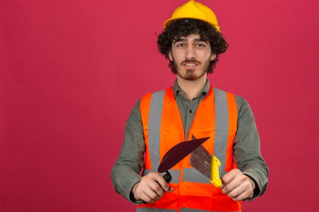 Photo gratuite jeune ingénieur beau barbu portant un casque de sécurité et un gilet tenant des couteaux à mastic avec sourire sur le visage sur un mur rose isolé