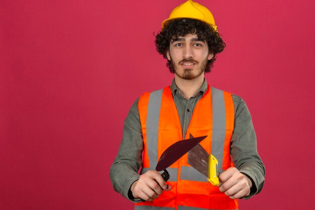 Jeune ingénieur beau barbu portant un casque de sécurité et un gilet tenant des couteaux à mastic avec sourire sur le visage sur un mur rose isolé