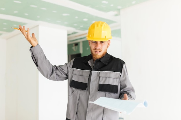 Jeune ingénieur émotionnel en vêtements de travail et casque jaune tenant le plan du nouvel appartement à la main regardant à huis clos au travail