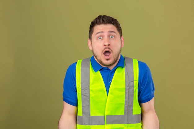 Jeune ingénieur homme portant un gilet de construction à fasciné par l'incrédulité surprise et expression étonnée debout sur fond vert isolé