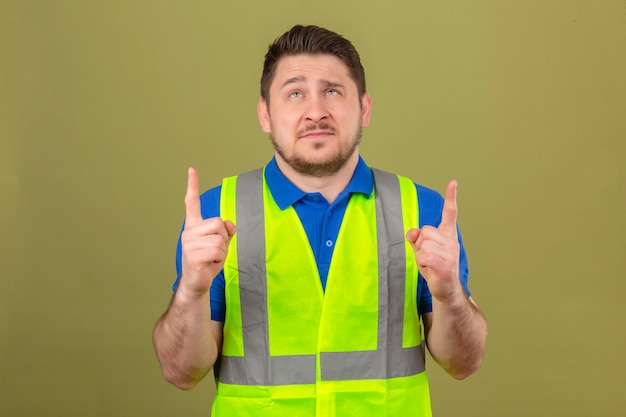 Photo gratuite jeune ingénieur homme portant un gilet de construction à la pointe des doigts vers le haut avec un sourire sceptique sur le visage sur fond vert isolé