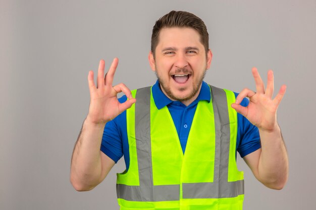 Jeune ingénieur homme portant un gilet de construction regardant la caméra avec un grand sourire et un visage heureux faisant des signes ok sur fond blanc isolé