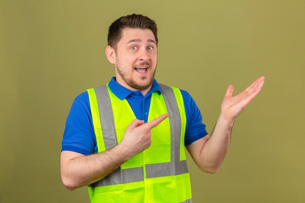 Jeune ingénieur homme portant un gilet de construction souriant à la caméra tout en présentant avec la main et en pointant avec le doigt sur fond vert isolé