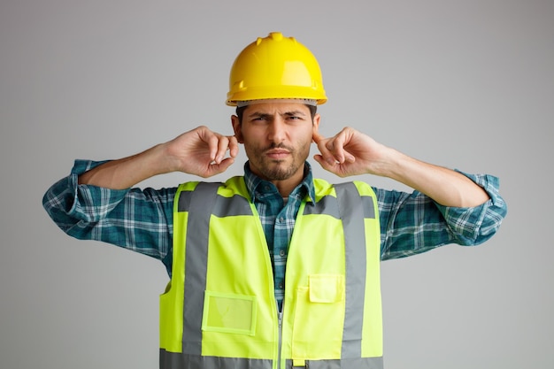 Jeune ingénieur mécontent portant un casque de sécurité et un uniforme regardant la caméra tout en gardant les doigts dans les oreilles isolés sur fond blanc