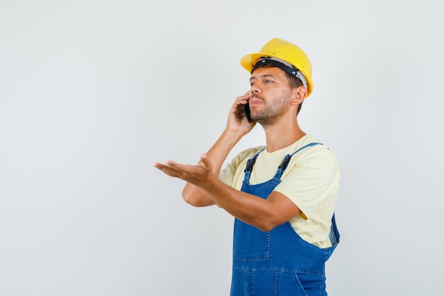 Jeune ingénieur parlant au téléphone avec la paume ouverte en uniforme et regardant pensif, vue de face.