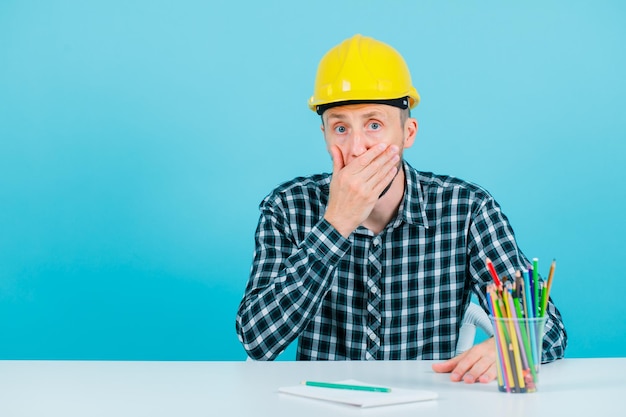 Un jeune ingénieur surpris couvre sa bouche avec la main sur fond bleu