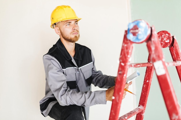 Jeune ingénieur en vêtements de travail et casque jaune regardant pensivement de côté s'appuyant sur une échelle rouge dans la réparation d'un appartement