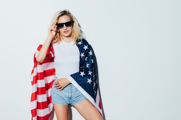 Jeune et insouciant. Belle jeune femme blonde portant le drapeau américain et souriant tout en célébrant le 4 juillet, jour de l'indépendance debout contre le mur blanc