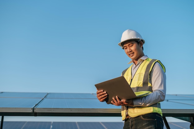 Jeune inspecteur asiatique Ingénieur homme utilise un ordinateur portable travaillant à la ferme solaire Technicien superviseur mâle en casque blanc Vérification du fonctionnement du soleil et du panneau solaire photovoltaïque dans l'espace de copie de la station