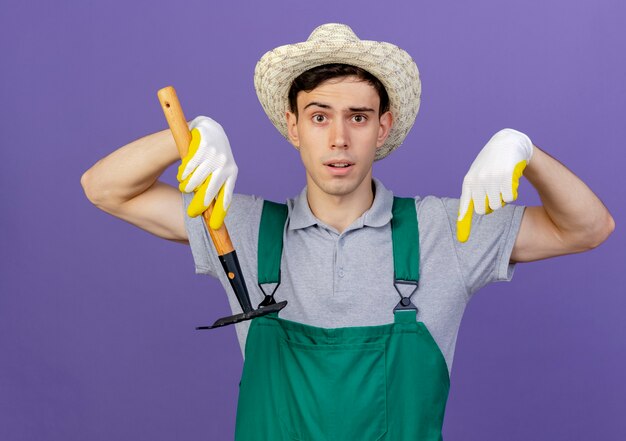 Jeune jardinier anxieux portant chapeau et gants de jardinage tient le râteau et pointe vers le bas avec deux mains