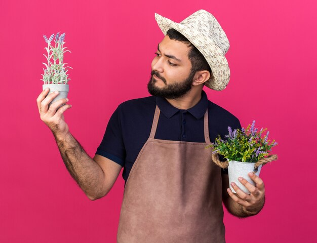 Jeune jardinier caucasien désemparé portant un chapeau de jardinage tenant des pots de fleurs isolés sur un mur rose avec espace pour copie