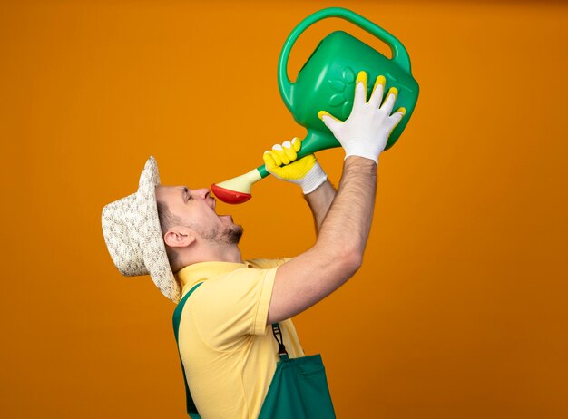 Jeune jardinier en combinaison et chapeau tenant un arrosoir essayant de boire de l'eau debout sur un mur orange