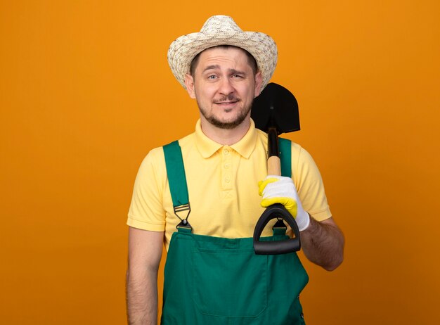 Jeune jardinier en combinaison et chapeau tenant une pelle à l'avant souriant confiant debout sur un mur orange