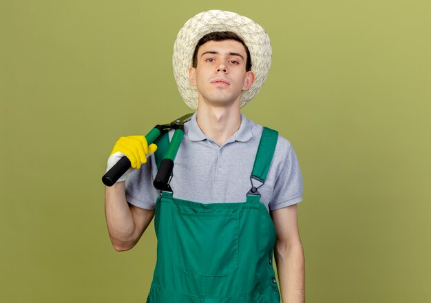 Un jeune jardinier confiant portant un chapeau et des gants de jardinage tient une tondeuse sur l'épaule