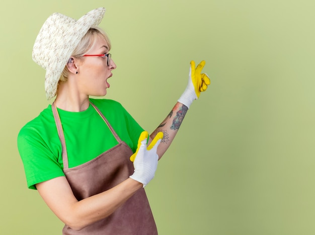 Jeune jardinier femme aux cheveux courts en tablier et chapeau portant des gants en caoutchouc pointant avec l'index à quelque chose en arrière à la surprise debout sur fond clair